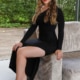 Leila - Escortmodel Stuttgart im langen schwarzen Kleid auf der Lehne eines Sofas sitzend.