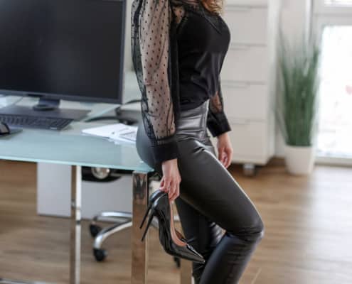 Angelina - Blondes Escort Dortmund in einer schwarzen Lederhose und schwarzer Bluse im Büro vor einem Tisch stehend.
