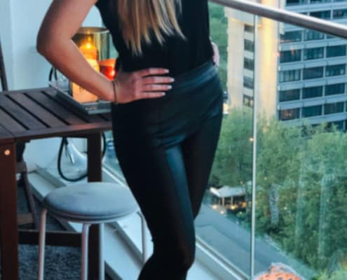 Alissa -charmantes Escort Düsseldorf in einer schwarzen Hose, einem engen Oberteil und Sandalen auf einem Balkon stehend.