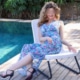 Angelina - Blondes Escort Dortmund im blauen Sommerkleid vor dem Pool auf einem Stuhl sitzend.