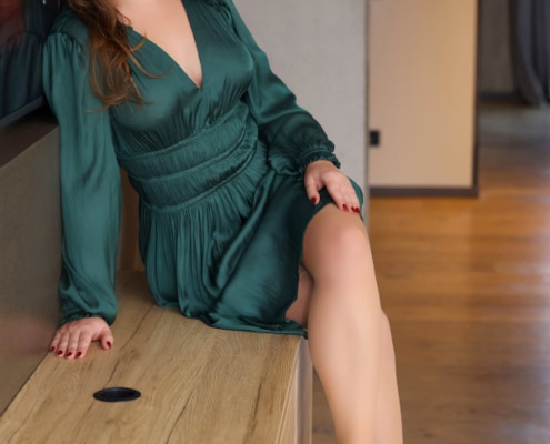 Sophia- VIP Escortmodel Berlin im grünen Kleid seitlich auf einer Kommode sitzend.