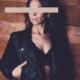 Gina - Escortmodel Köln im schwarzen BH und Lederjacke vor einer Holzwand stehend.
