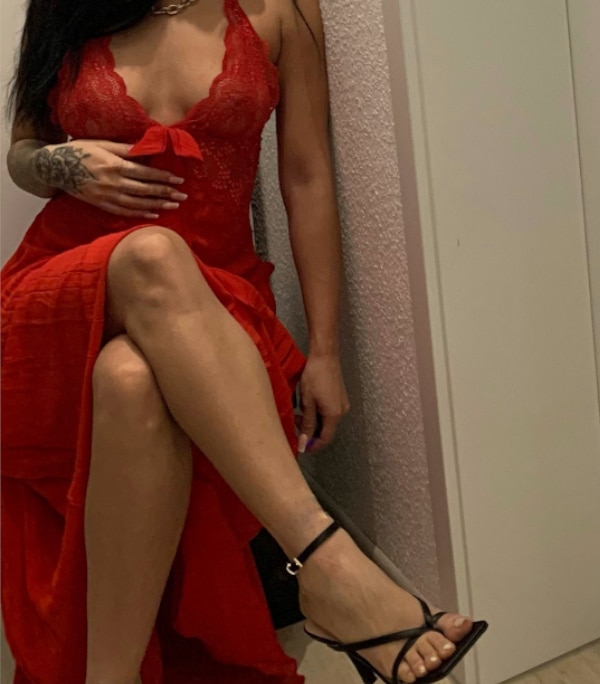 Alessia - Escort Heilbronn in einem engen roten Kleid mit schwarzen Sandalen vor einem Spiegel sitzend.