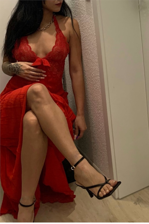 Alessia - Escort Heilbronn in einem engen roten Kleid mit schwarzen Sandalen vor einem Spiegel sitzend.