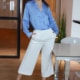 Luisa - Escort Bamberg in blauer Bluse und weisser Hose vor einem Glastisch stehend.