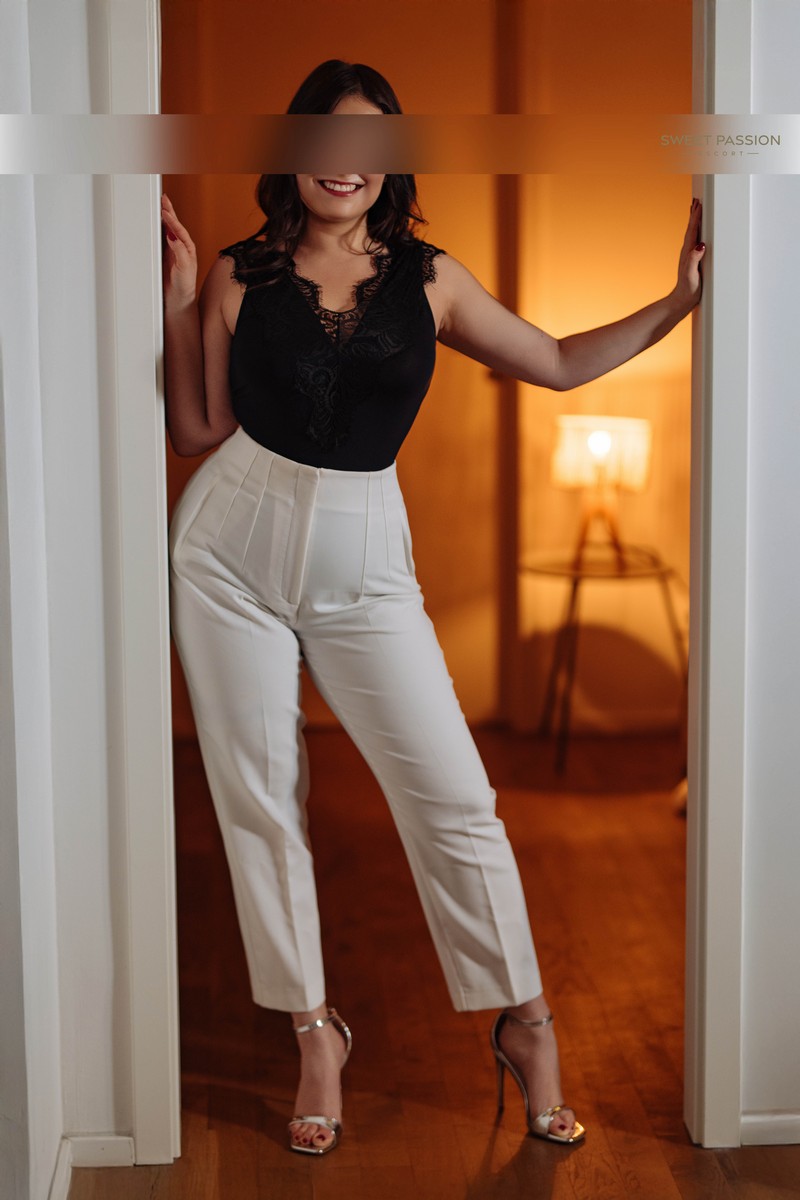 malina - curviges escort muenchen im schwarzen Top, weisser Anzughose und goldenen Highheel Sandalen. Sie steht im Türrahmen und lächelt.